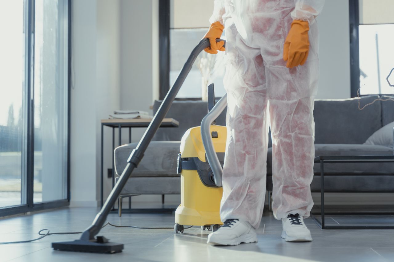 services-02 A cleaner in protective coveralls and gloves vacuuming a modern living room.