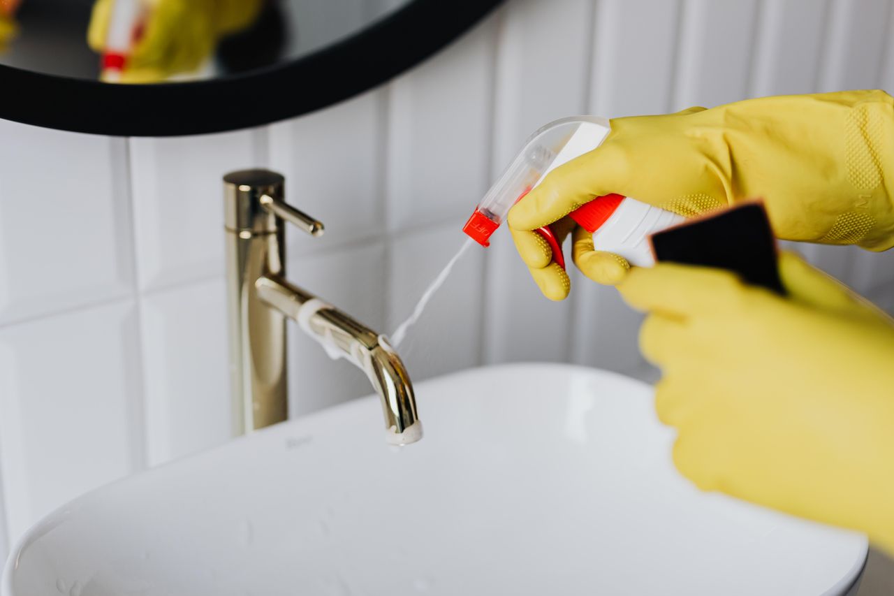 heros-img From above anonymous housekeeper in yellow rubber gloves spraying cleaner to chrome water faucet in bathroom