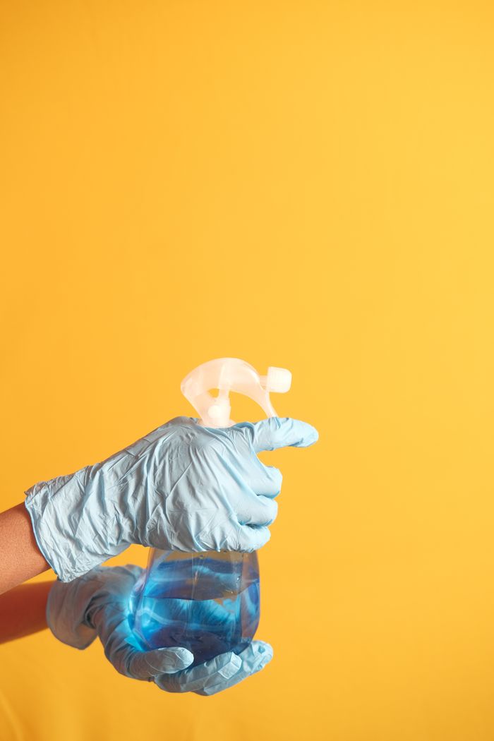 about-us-01 Hand in blue glove holding a cleaning spray bottle against a yellow background.